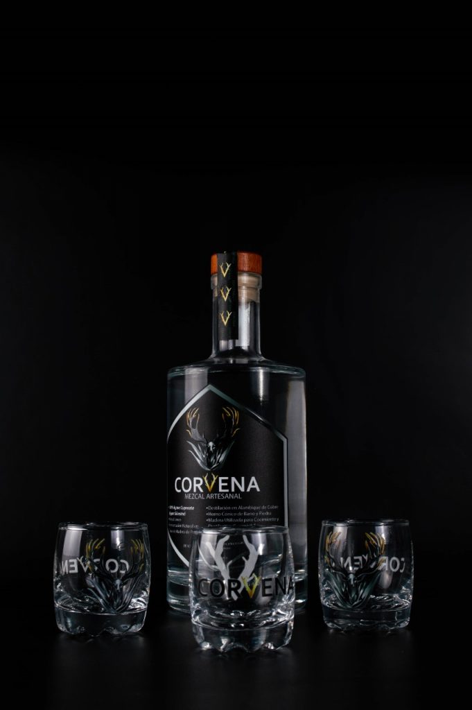 Botella de Mezcal Artesanal Corvena con tres vasos tequileros serigrafiados sobre un fondo negro sólido.