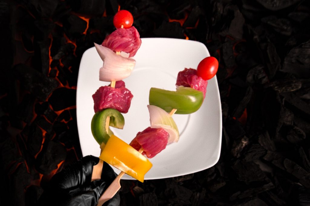Mano con guante negro sosteniendo brochetas de carne cruda, pimientos y cebolla sobre un plato blanco y fondo de carbón encendido.