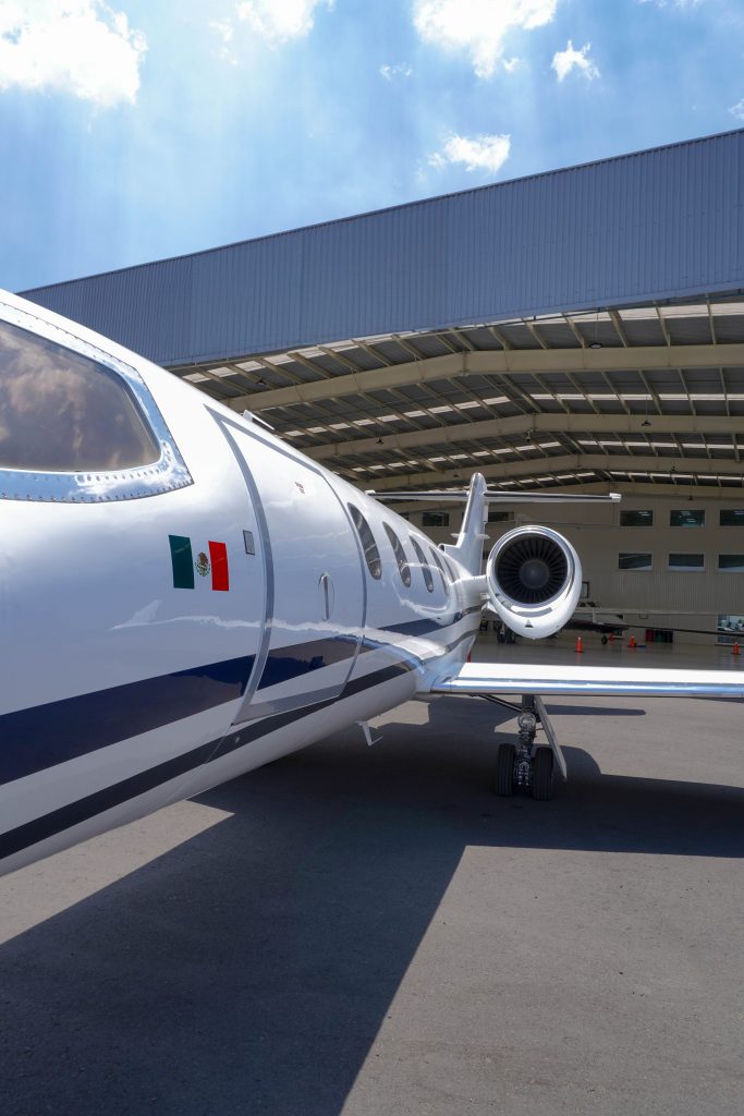 Primer plano lateral de un jet privado blanco mostrando la bandera de México y la turbina del motor frente a un hangar.