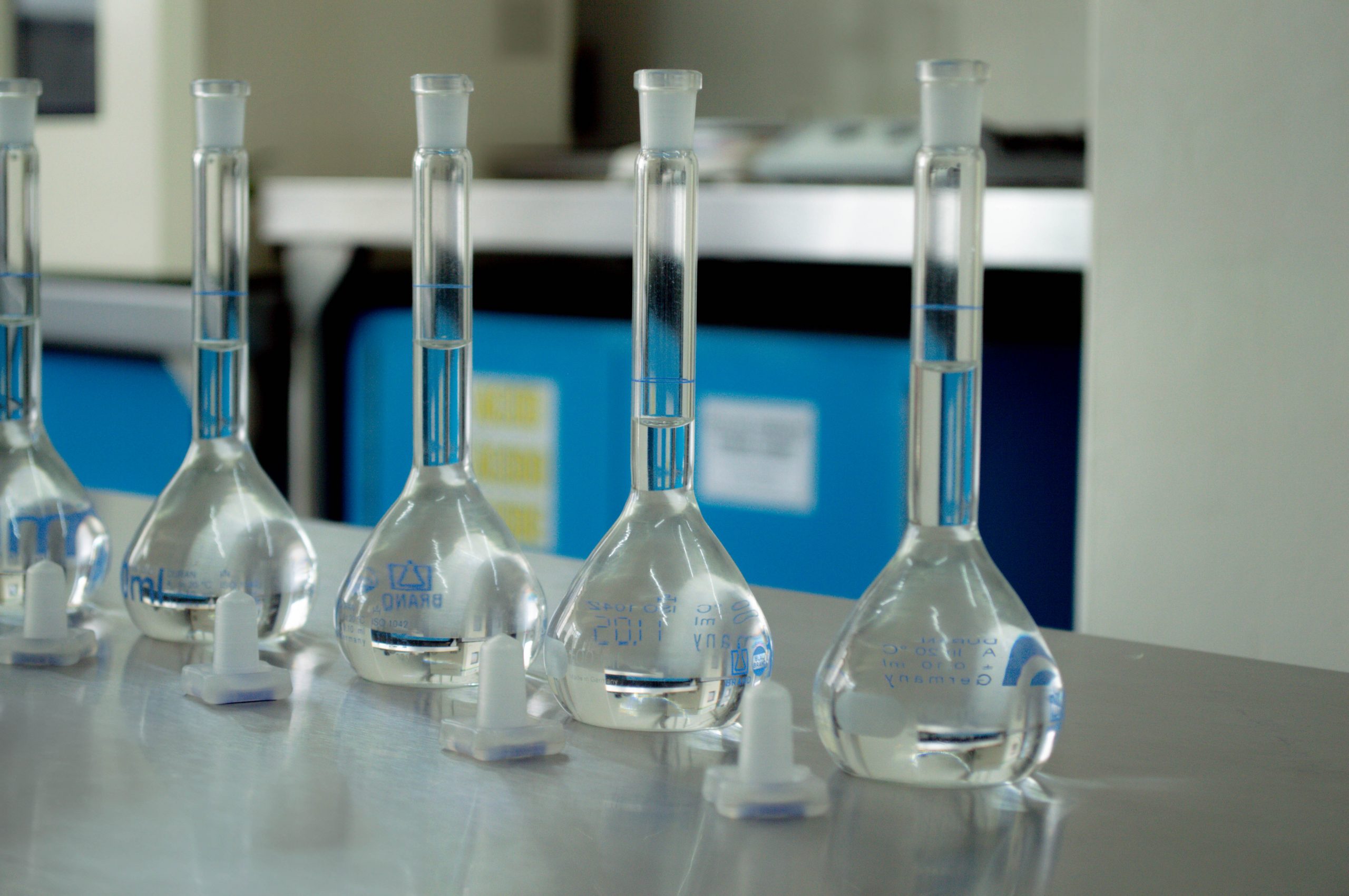 Fila de matraces aforados de vidrio con líquido transparente sobre una mesa de acero inoxidable en un laboratorio químico profesional.